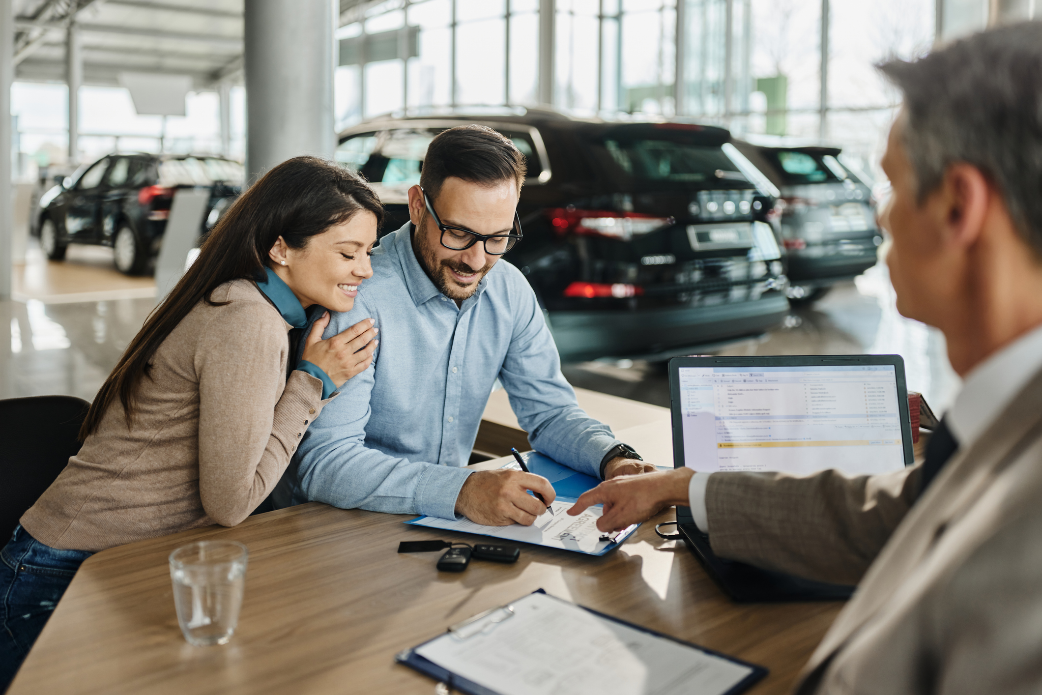 Couple sitting at a desk in a car dealership signing a paper.