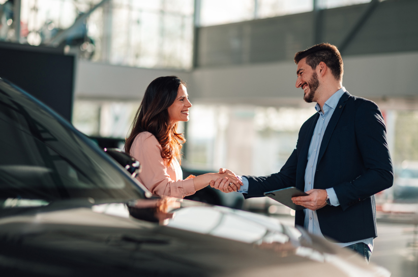 Car dealer shaking hands with a woman buyer in front of a vehicle.