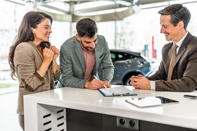 Couple signing a paper standing at the car dealership while the car dealer watches them happily.