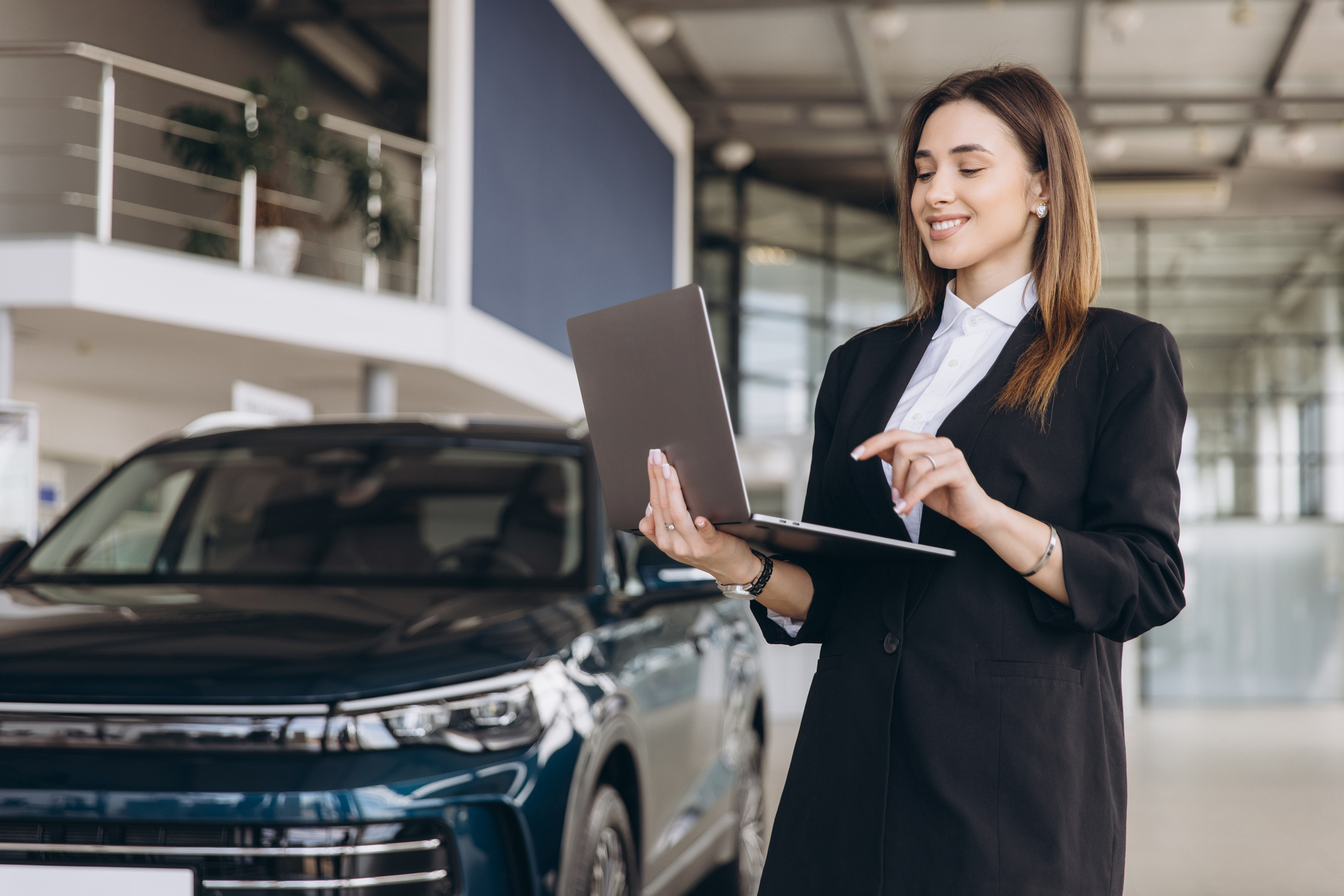 Saleswoman standing next to a new car in a dealership, using a laptop to assist customers with online vehicle purchases