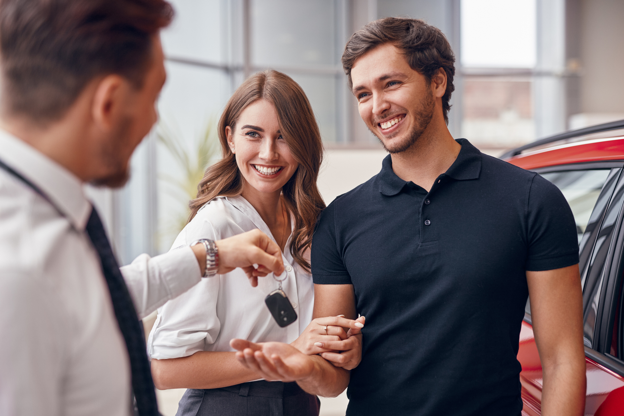 Blurred male dealer giving key of new car to excited young couple in modern auto salon.