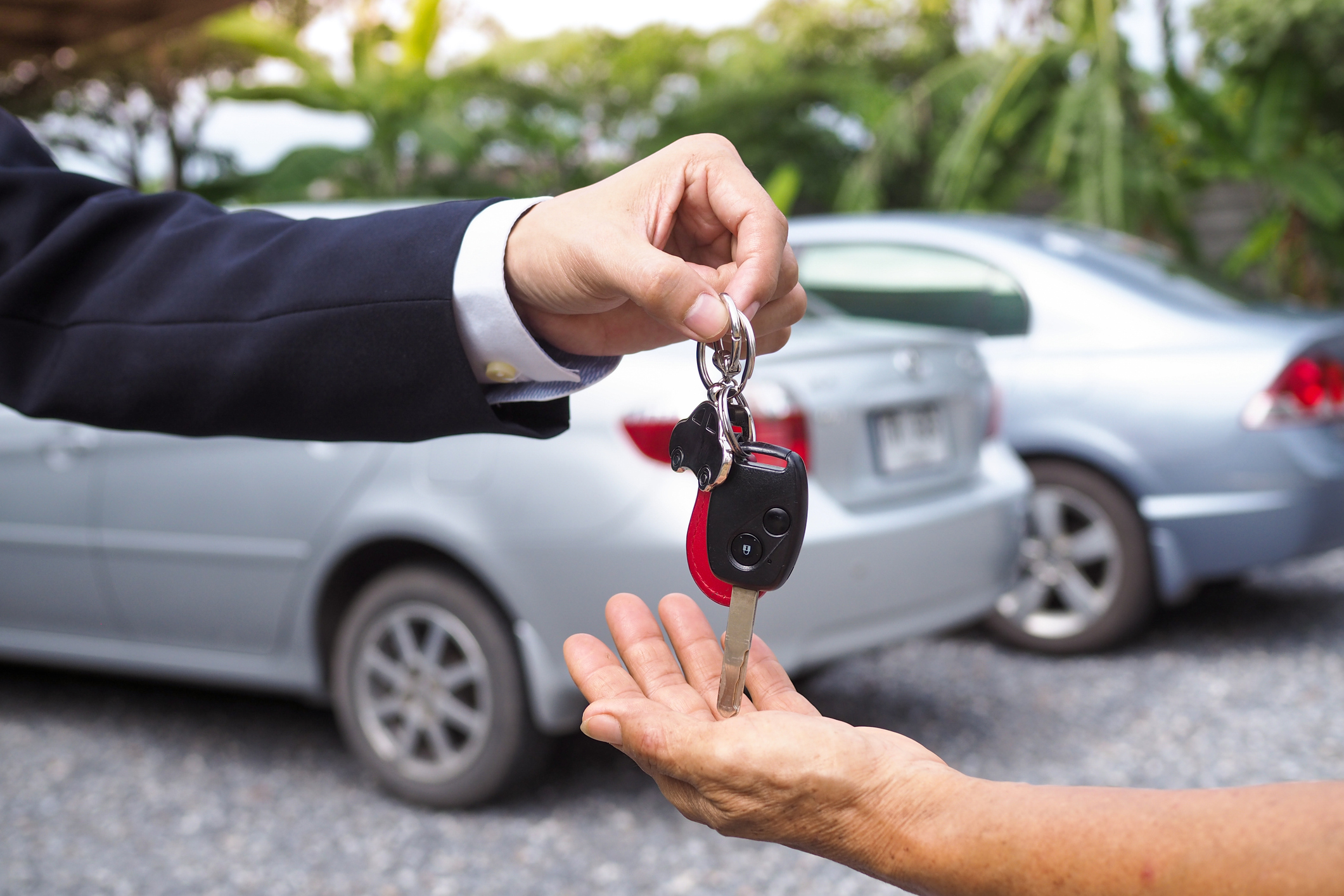 A person handing keys to a car buyer in front of two gray cars outside.