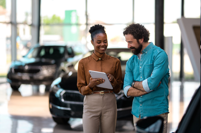 Two people looking at a clipboard in the middle of a car dealership.