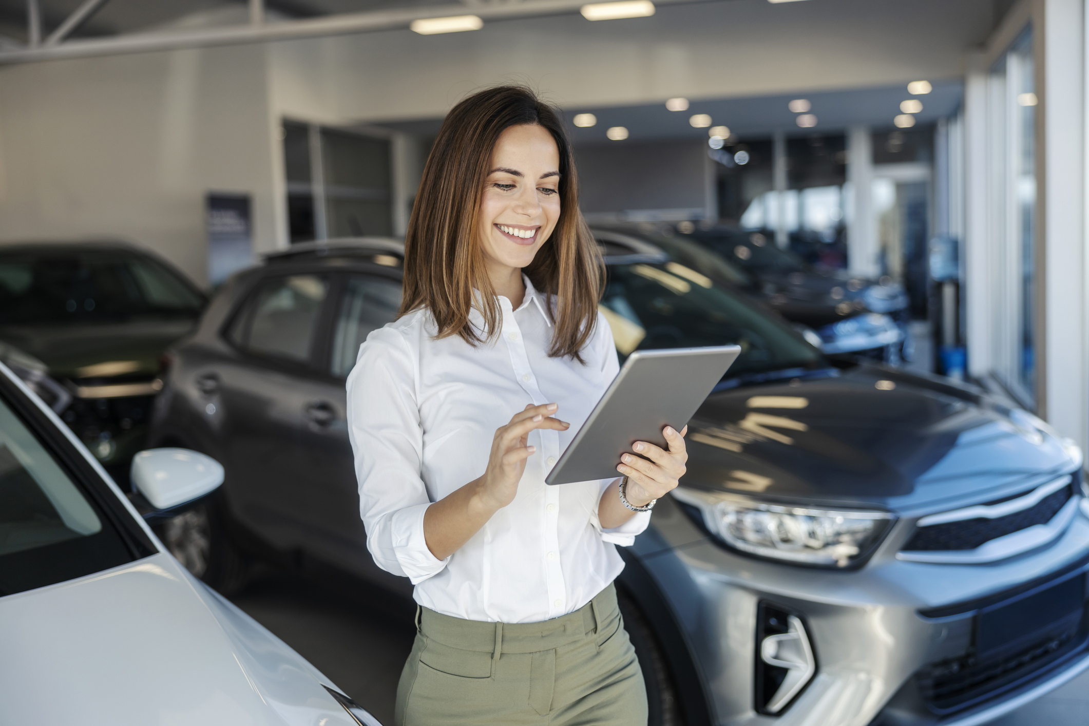 female car dealer standing at car showroom with tablet in hands and scrolling on it.